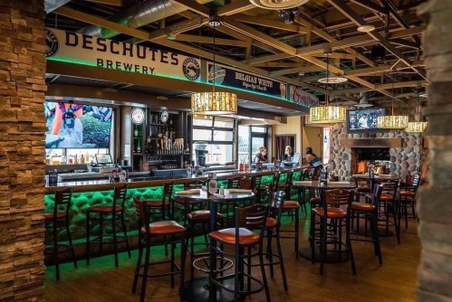 Interior of a modern brewery with a bar, high stools, and a cozy fireplace, featuring green lighting and multiple TVs.