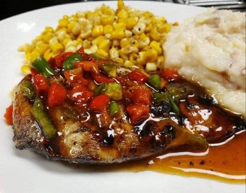 Grilled chicken topped with colorful peppers and sauce, served with corn and mashed potatoes on a white plate.