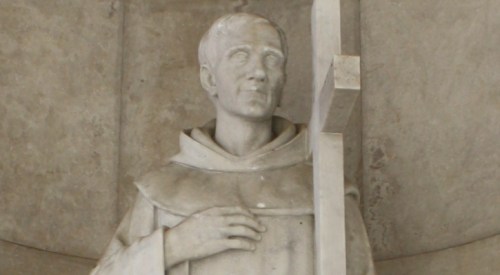A marble statue of a monk holding a cross, with a contemplative expression, set against a textured background.