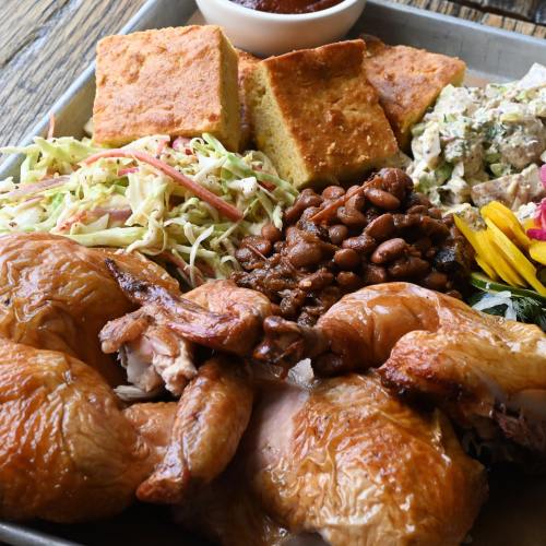 A platter featuring roasted chicken, cornbread, coleslaw, baked beans, and a side salad, garnished with flowers.