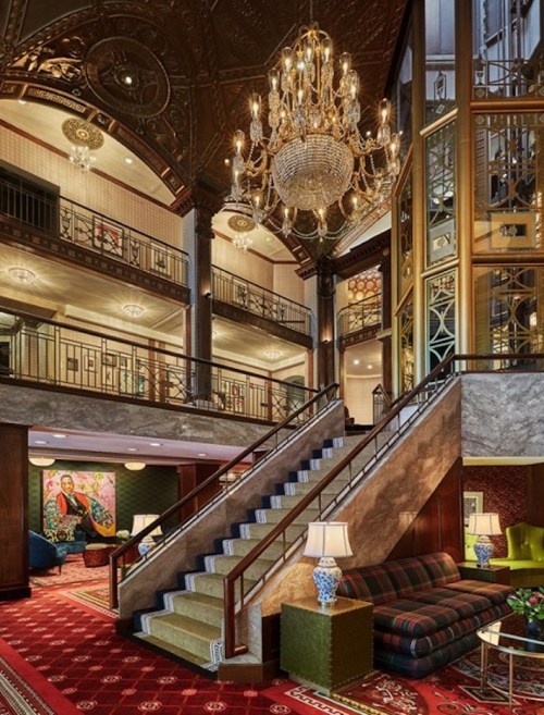 Elegant hotel lobby featuring a grand staircase, chandelier, and stylish seating areas with rich decor.