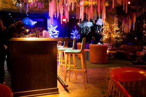 A cozy bar scene with colorful stools, ambient lighting, and floral decorations hanging from the ceiling.