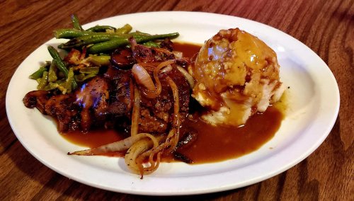 A plate of grilled chicken with onions, green beans, and mashed potatoes topped with gravy.