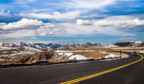 A winding road through a mountainous landscape with snow-capped peaks and a blue sky dotted with clouds.