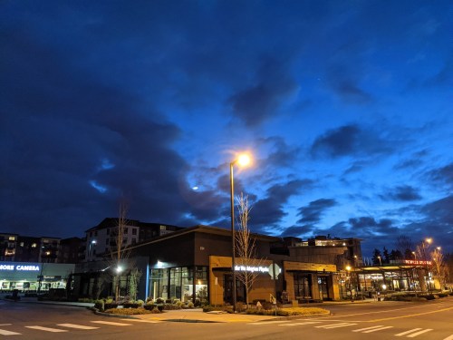 A dark blue sky with clouds above a street corner featuring illuminated buildings and a streetlight.