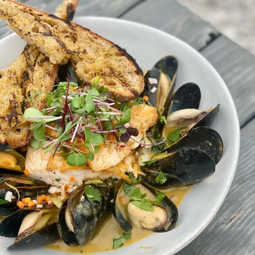 A plate of grilled fish topped with microgreens, surrounded by mussels and served with toasted bread.