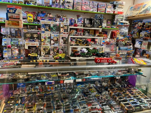 A cluttered display of toys and collectibles, including cars, action figures, and various merchandise on shelves and counters.