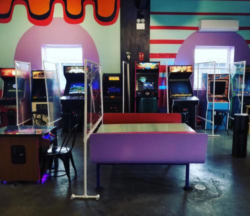 A colorful arcade with retro game machines, separated by clear barriers, and a purple seating area in the center.