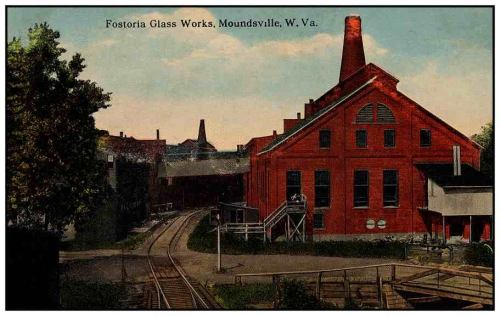 Historic Fostoria Glass Works building in Moundsville, West Virginia, with smokestacks and railway tracks.
