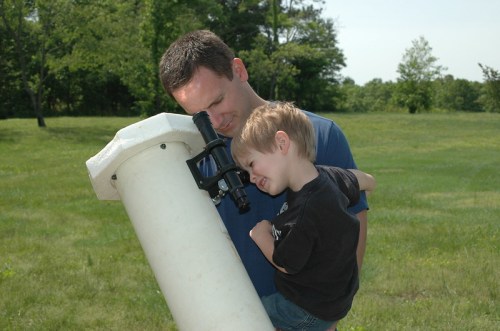 A man and a young boy look through a telescope in a grassy field on a sunny day.