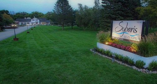 A well-maintained lawn leads to a sign for Sybaris Pool Suites, with a house and trees in the background at dusk.