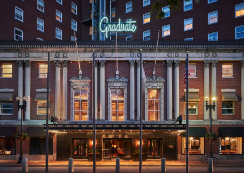 The exterior of the Graduate hotel, featuring classic architecture and a neon sign, with flags and street lamps.
