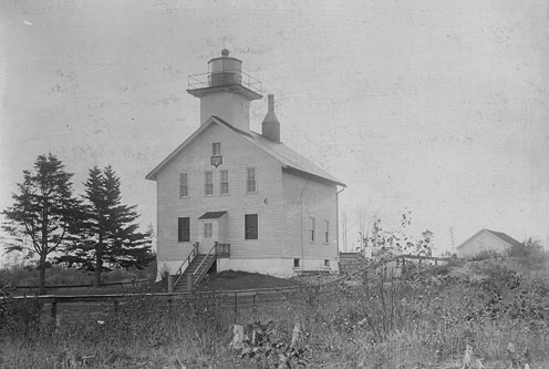 Historic lighthouse with a white wooden structure, surrounded by trees and a grassy area, under a cloudy sky.