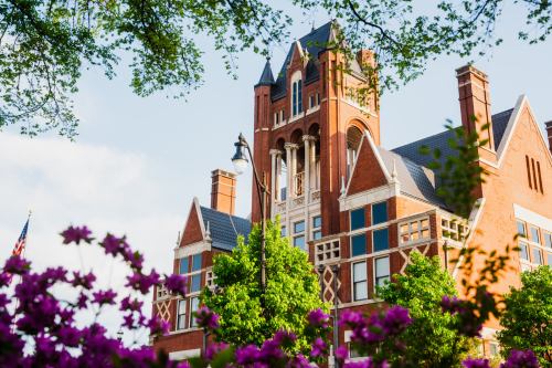 Historic brick building with a tower, surrounded by green trees and blooming purple flowers under a clear sky.