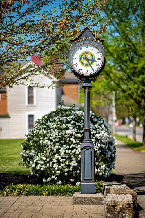 A decorative clock stands on a sidewalk, surrounded by blooming white flowers and greenery in a sunny setting.
