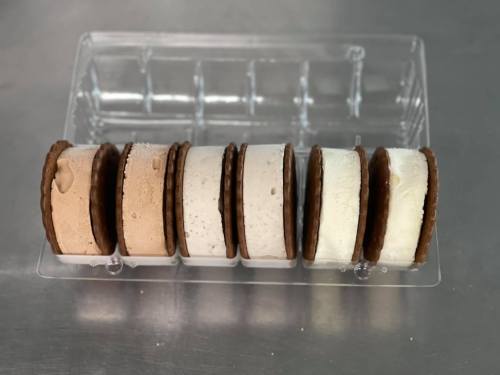 A clear container holding six ice cream sandwiches with chocolate cookies and various ice cream flavors.