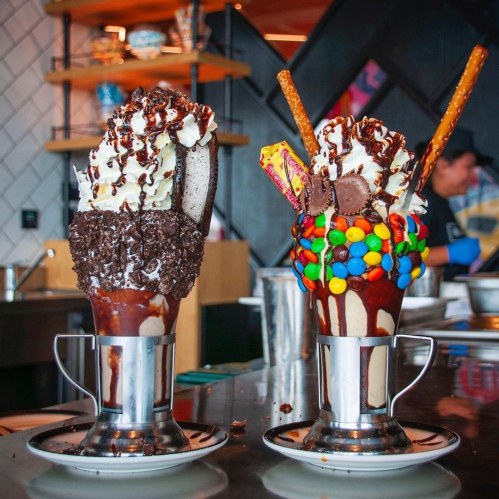 Two extravagant milkshakes topped with whipped cream, chocolate drizzle, and colorful candies, served in metal cups.