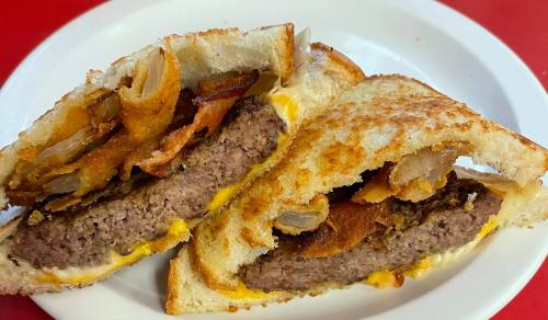 A close-up of a grilled sandwich filled with a beef patty, cheese, crispy bacon, and fried onions on toasted bread.
