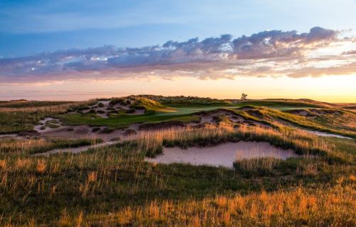 A scenic golf course at sunset, featuring rolling hills, sand traps, and a colorful sky with clouds.