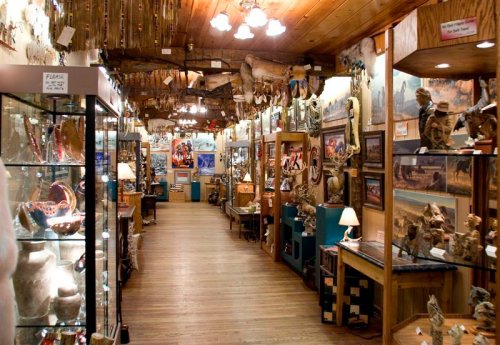 A spacious art gallery filled with various sculptures, paintings, and decorative items, featuring wooden beams and warm lighting.