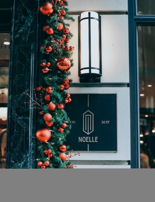 A festive entrance with a garland of greenery and red ornaments, featuring a sign that reads "Noelle" and the years 1930-2017.