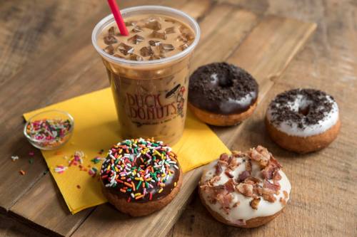 A cup of iced coffee next to three donuts: one with chocolate icing and sprinkles, one with bacon, and one with crushed cookies.