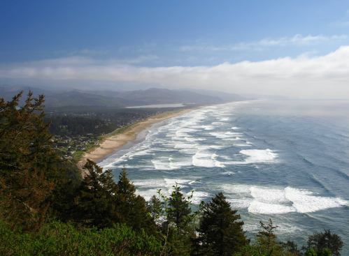 A scenic view of a coastline with waves crashing on the shore, surrounded by lush greenery and distant mountains.