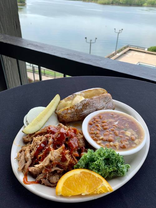 A plate of pulled pork with barbecue sauce, baked potato, beans, kale, and an orange slice, with a lake view in the background.