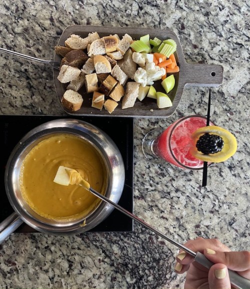 A hand holding a fondue fork with cheese, alongside a platter of bread and veggies, and a colorful drink with a garnish.