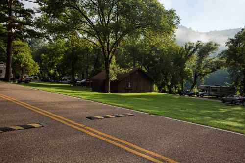A peaceful campsite with a wooden cabin, surrounded by trees and a misty mountain backdrop, along a paved road.