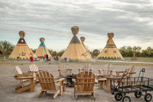 A circle of wooden chairs surrounds a fire pit, with colorful tipis in the background under a cloudy sky.