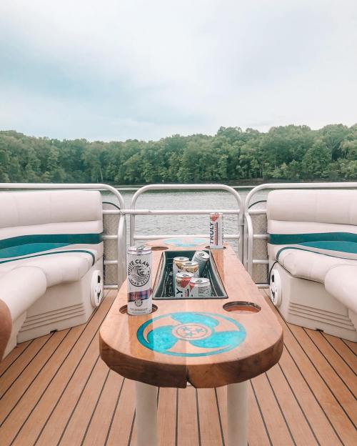 A boat's deck with a wooden table holding drinks, surrounded by green trees and calm water in the background.