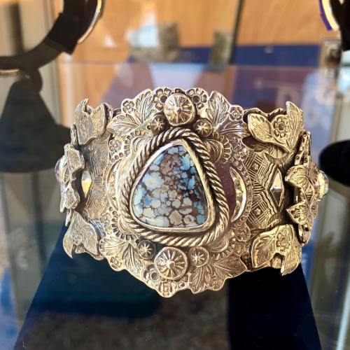 A detailed silver cuff bracelet featuring intricate floral designs and a triangular turquoise stone centerpiece.