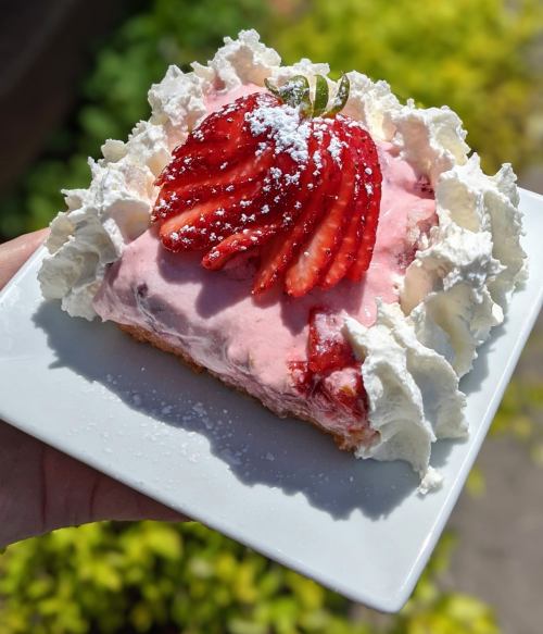 A slice of strawberry dessert topped with whipped cream and fresh strawberries, served on a white plate.