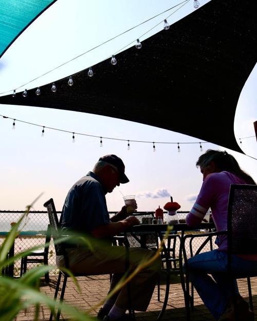 A couple sits at a table under a shaded area, enjoying drinks with string lights overhead and a scenic view in the background.