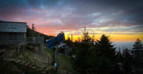 A scenic view of a sunset behind wooden cabins and trees, with vibrant colors in the sky.