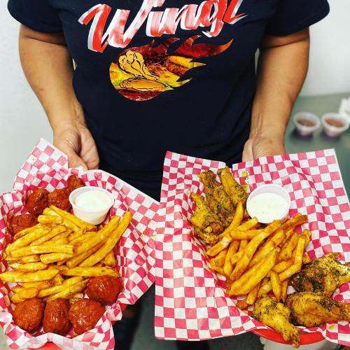 A person holds two trays of food: one with chicken wings and fries, the other with meatballs and fries, both in checkered baskets.