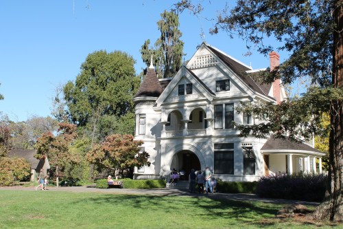 A large, elegant white house with a manicured lawn, surrounded by trees and people enjoying the outdoor space.
