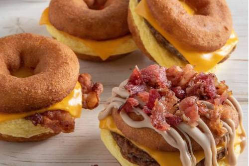 Three savory breakfast sandwiches featuring donuts, cheese, eggs, sausage, and crispy bacon on a wooden surface.