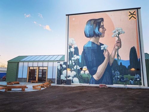 A large mural of a woman holding flowers on a building, with a modern structure and outdoor seating area nearby.