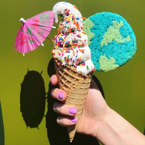 A hand holding an ice cream cone topped with sprinkles, a cookie shaped like the Earth, and a pink umbrella.