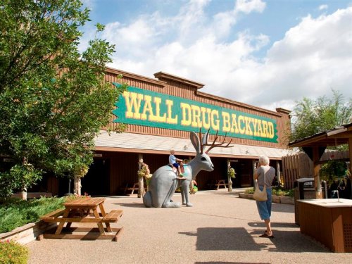 A large sign reading "Wall Drug Backyard" with visitors near a giant metal deer sculpture and picnic tables.