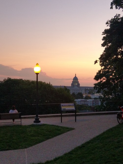 A sunset view over a city, featuring a lamppost and a distant dome building, with trees framing the scene.