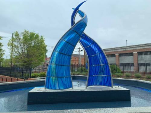 A modern blue glass fountain with two curved structures, surrounded by a small pool and greenery in the background.