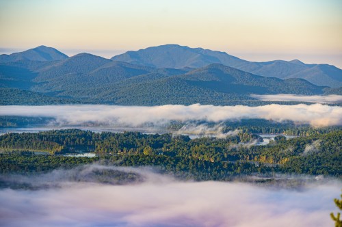 A panoramic view of misty mountains and valleys, with lush forests and a river winding through the landscape at sunrise.