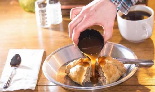 A hand pours sauce over a split pastry filled with meat, accompanied by a spoon and a cup of coffee on a wooden table.