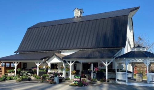 A large white barn with a black roof, featuring a sign for pies and surrounded by flowers and greenery.