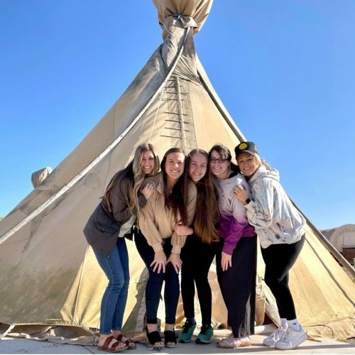 Five women pose together in front of a large tent under a clear blue sky, smiling and enjoying their time.