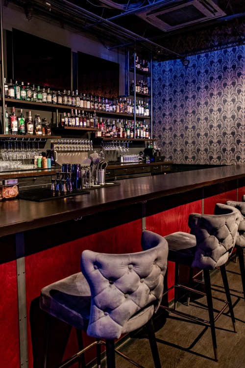 A stylish bar with a sleek countertop, plush gray stools, and a well-stocked shelf of liquor bottles in a dimly lit setting.