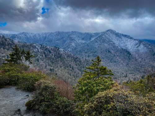 A scenic view of snow-dusted mountains under a cloudy sky, surrounded by lush greenery.